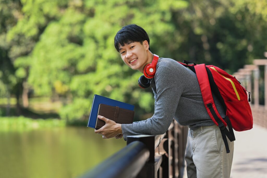 Portrait of smiling young college student with books and backpack standing on bridge over a river