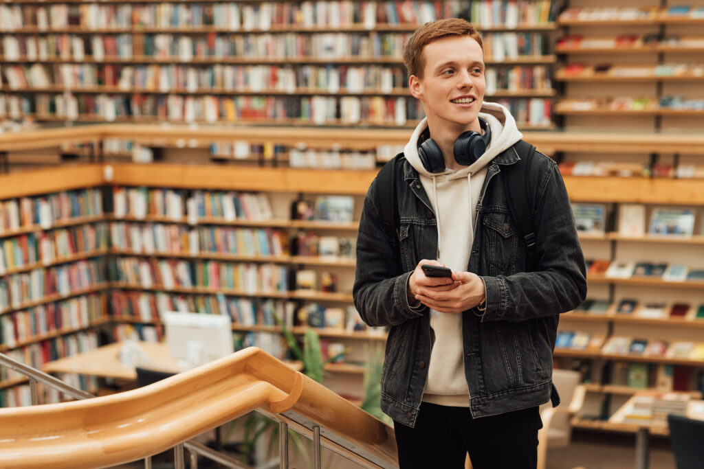 Smiling student with headphones and smartphone standing in university library